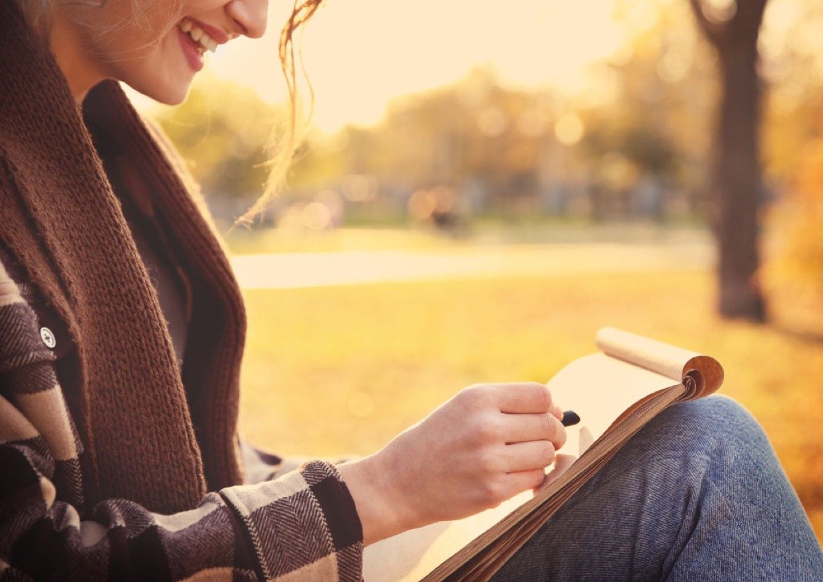 A person sitting outdoors in golden autumn light, smiling as they sketch in a book — a moment of flow